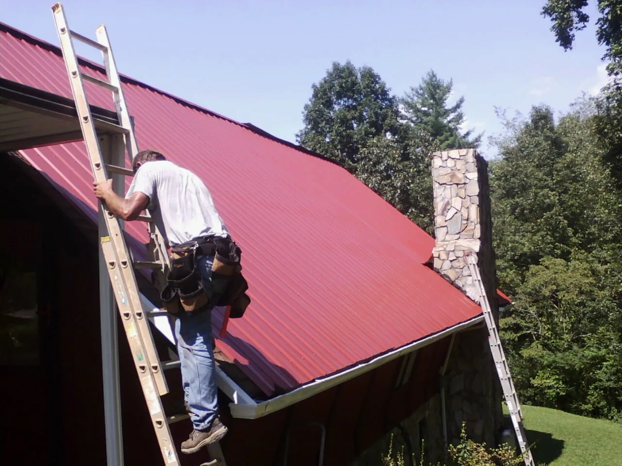 Roofer on a ladder beside a red metal roof and stone chimney — Soffit & Fascia Repair by Roofing Brothers in Rancocas